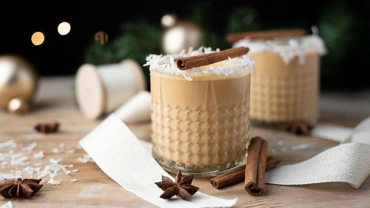 Two glasses of creamy drink topped with shredded coconut and cinnamon sticks, placed on a wooden surface with star anise, ribbon, and festive decorations in the background.