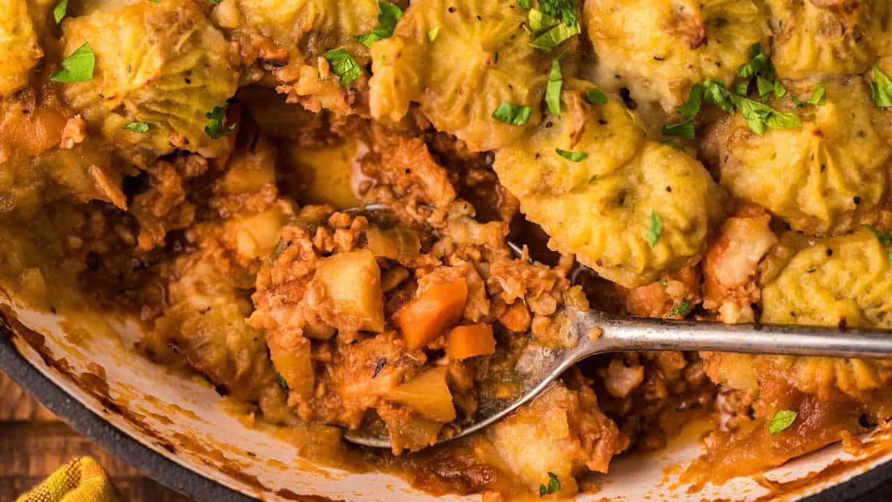 Close-up of a spoon scooping a portion of shepherd's pie showing ground meat, vegetables, and piped mashed potato topping, garnished with chopped parsley.