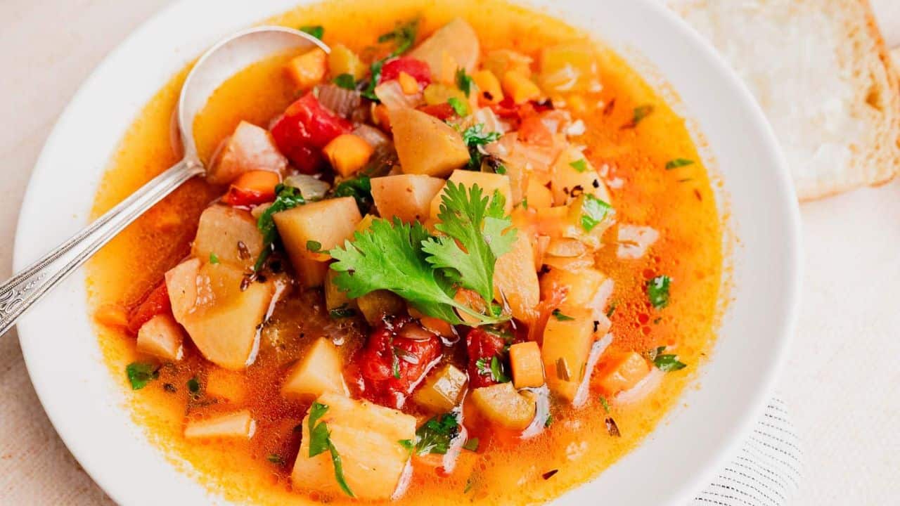 A white bowl filled with vegetable soup containing potatoes, carrots, tomatoes, and herbs, garnished with cilantro, with a spoon and a slice of bread on the side.