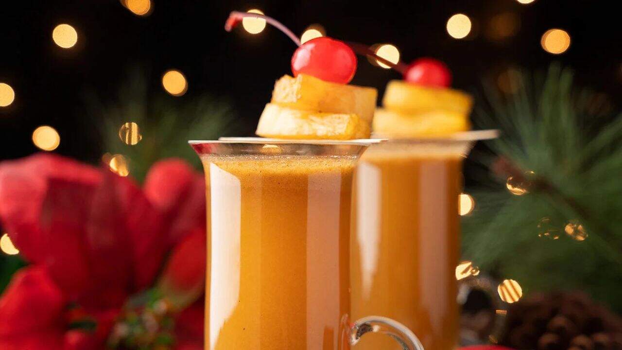 Two glasses of a frothy orange beverage garnished with pineapple slices and a maraschino cherry, with festive lights and holiday decor in the background.