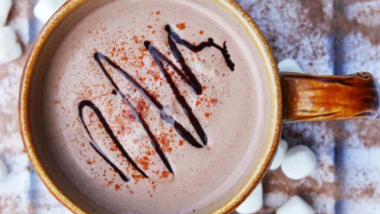 A brown ceramic mug filled with hot chocolate, topped with chocolate drizzle and cocoa powder, sits on a wooden surface with mini marshmallows scattered nearby.