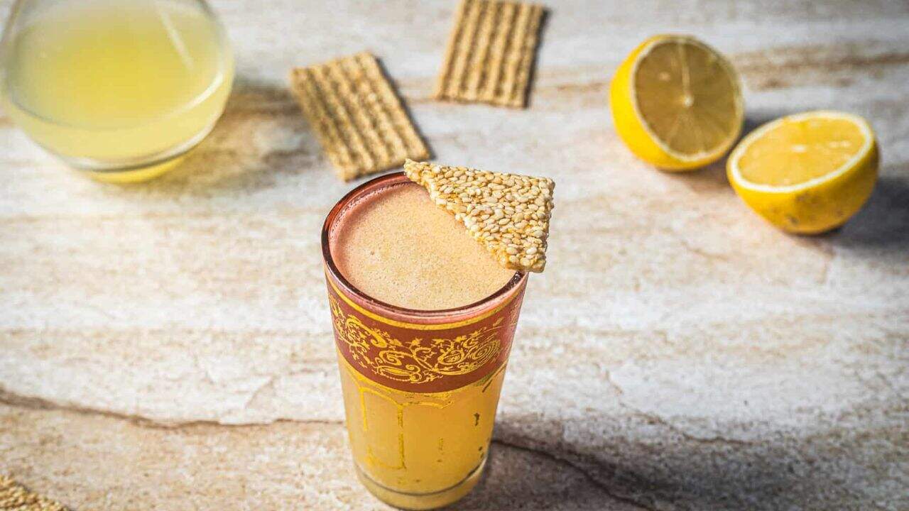 A glass of frothy yellow beverage garnished with sesame seeds and topped with a triangular piece of sesame brittle. A gold spoon, a lemon half, and another glass are in the blurred background.