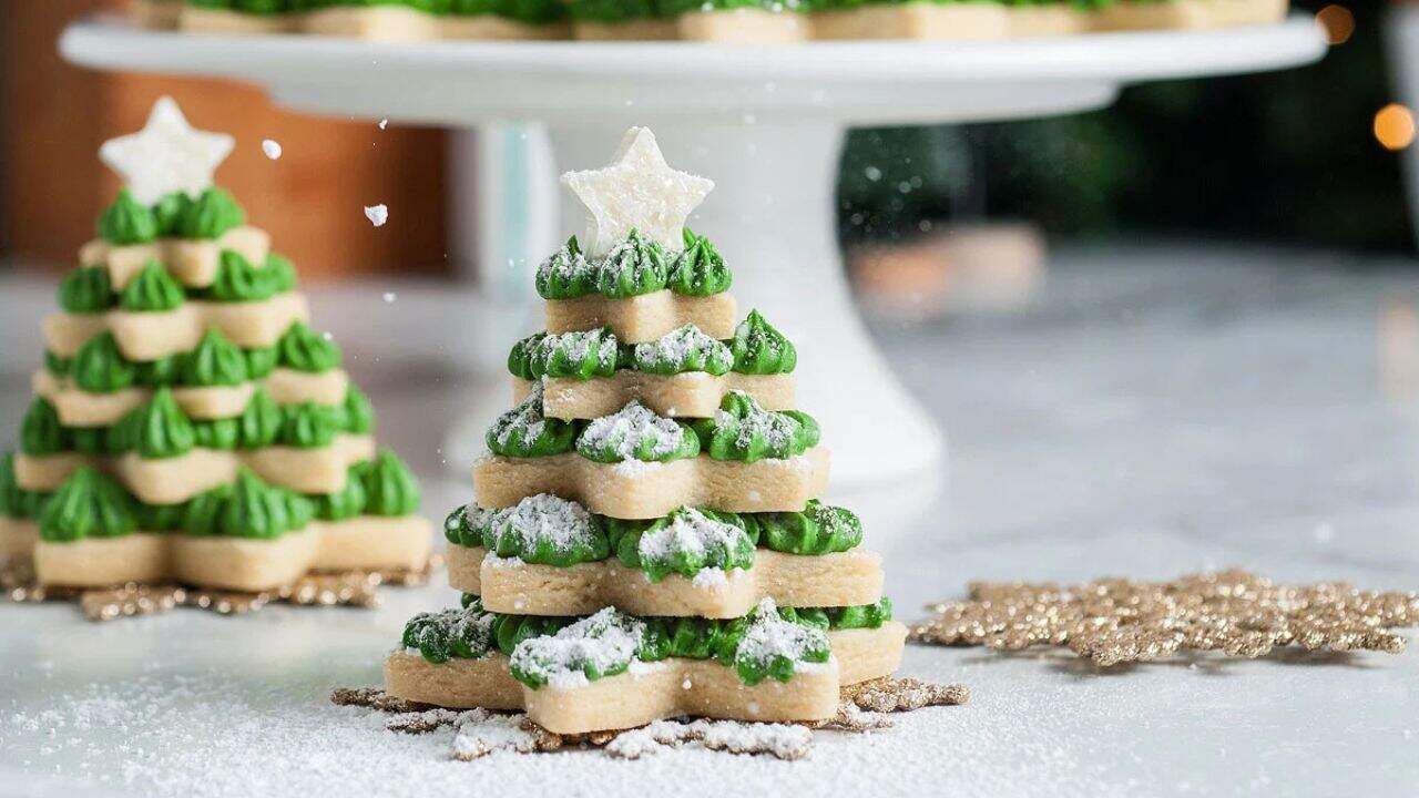 Stacked sugar cookies with green frosting form a Christmas tree shape, topped with a star cookie and dusted with powdered sugar, on a marble surface.