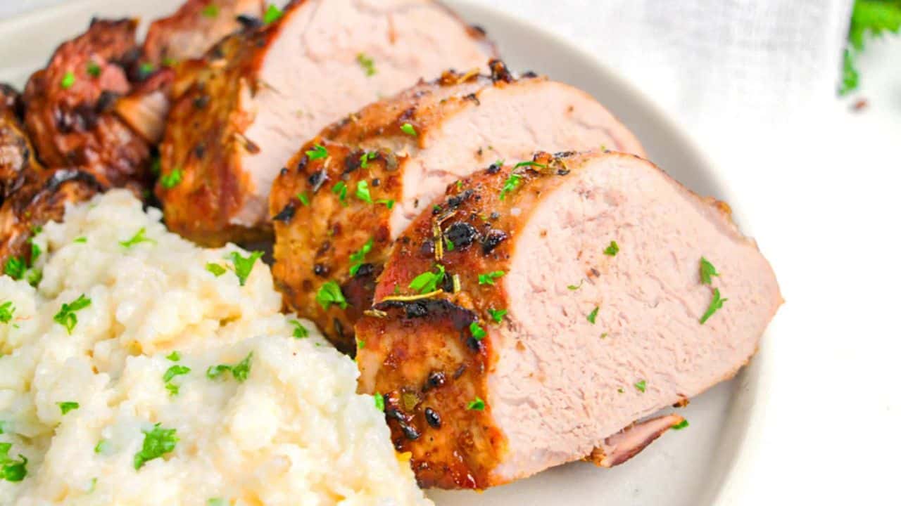 Sliced roasted pork tenderloin garnished with herbs, served next to a portion of mashed potatoes on a white plate.