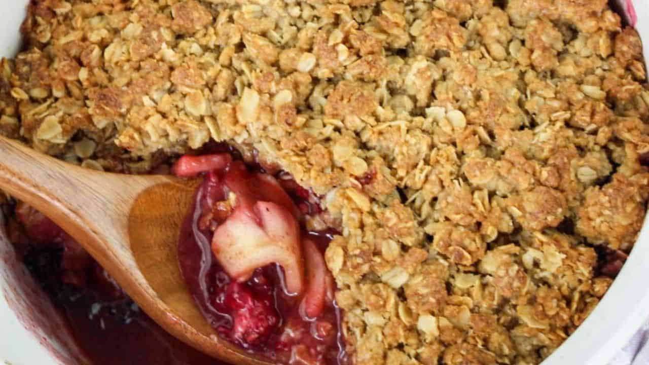 A bowl of fruit crumble with oats on top, featuring apples and blackberries. A fresh apple and blackberries are placed beside the bowl.