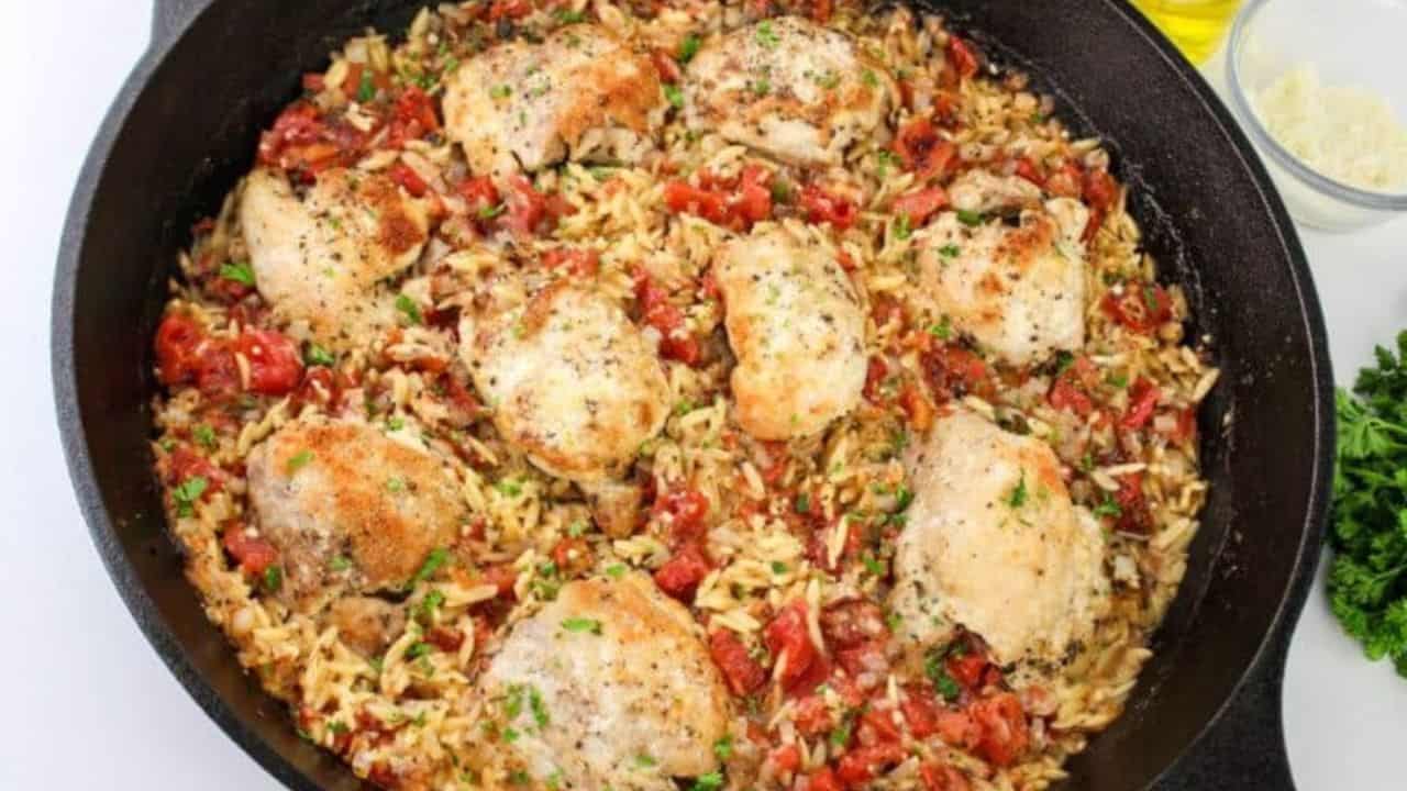 A plate with baked chicken thigh and orzo pasta mixed with tomatoes, garnished with herbs, with a fork and knife beside.