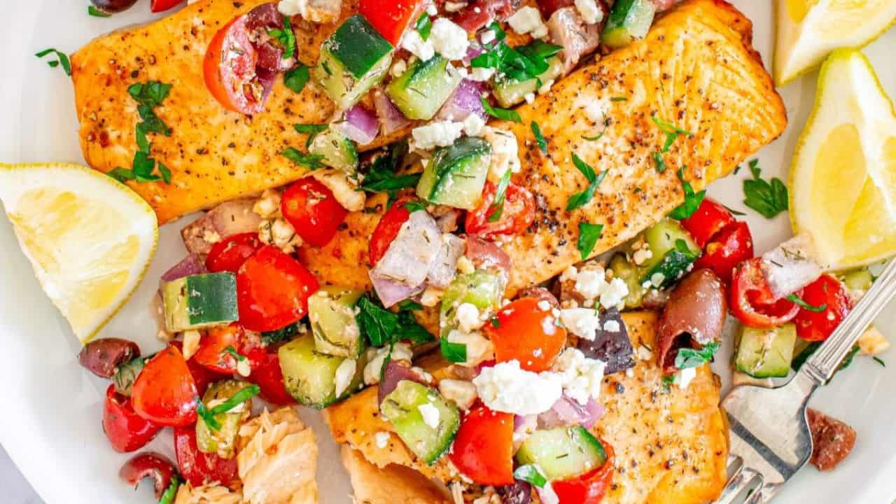 Grilled salmon fillets topped with a Mediterranean salad of tomatoes, cucumbers, olives, onions, and feta cheese, served with lemon wedges on a white plate.