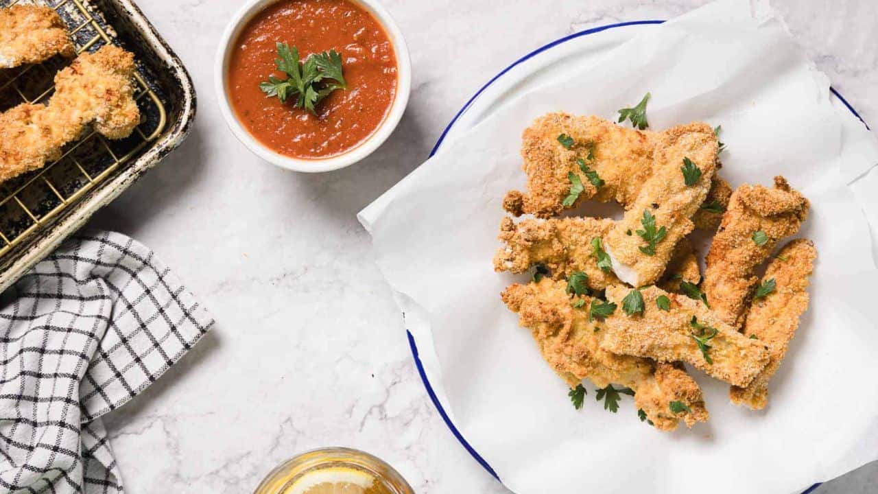 A plate of breaded chicken tenders is arranged around a small bowl of red dipping sauce, garnished with a sprig of parsley, on a paper-lined plate.