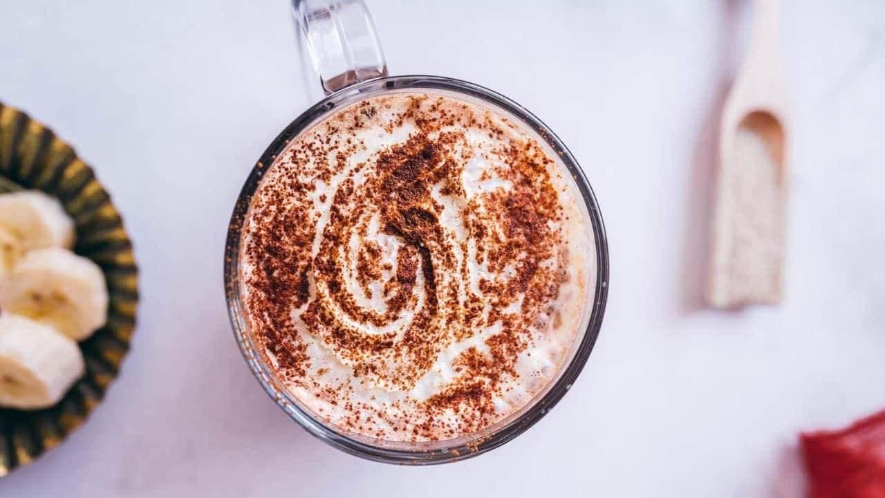 A glass mug filled with a creamy beverage topped with whipped cream and sprinkled cinnamon, viewed from above; banana slices and a wooden utensil are nearby on the table.
