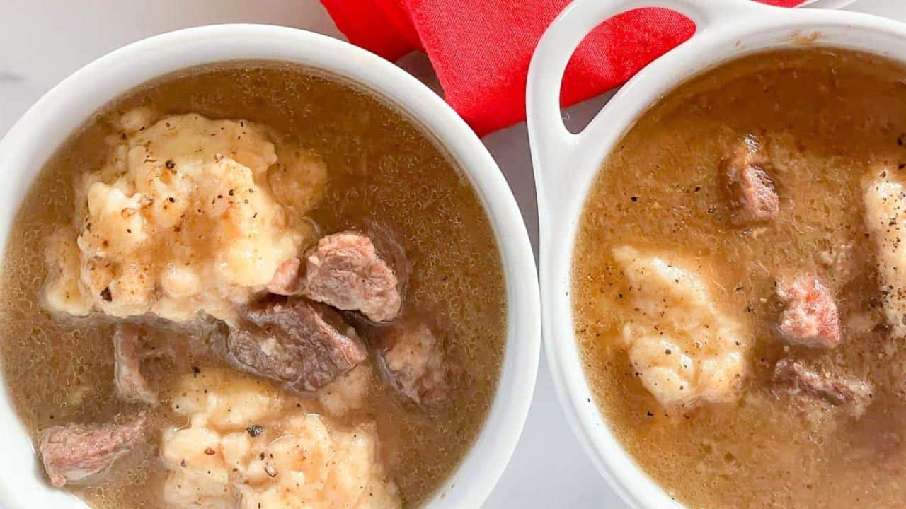Two bowls of beef stew with dumplings on a white surface. A red napkin and a spoon with salt are beside them.