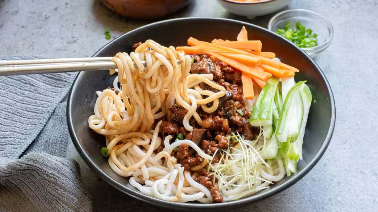 A bowl of noodles with sauce, garnished with sliced cucumbers, carrots, and bean sprouts. Using chopsticks, someone is lifting a portion of noodles. The dish is served in a white bowl, set on a light-colored table with a textured napkin nearby.