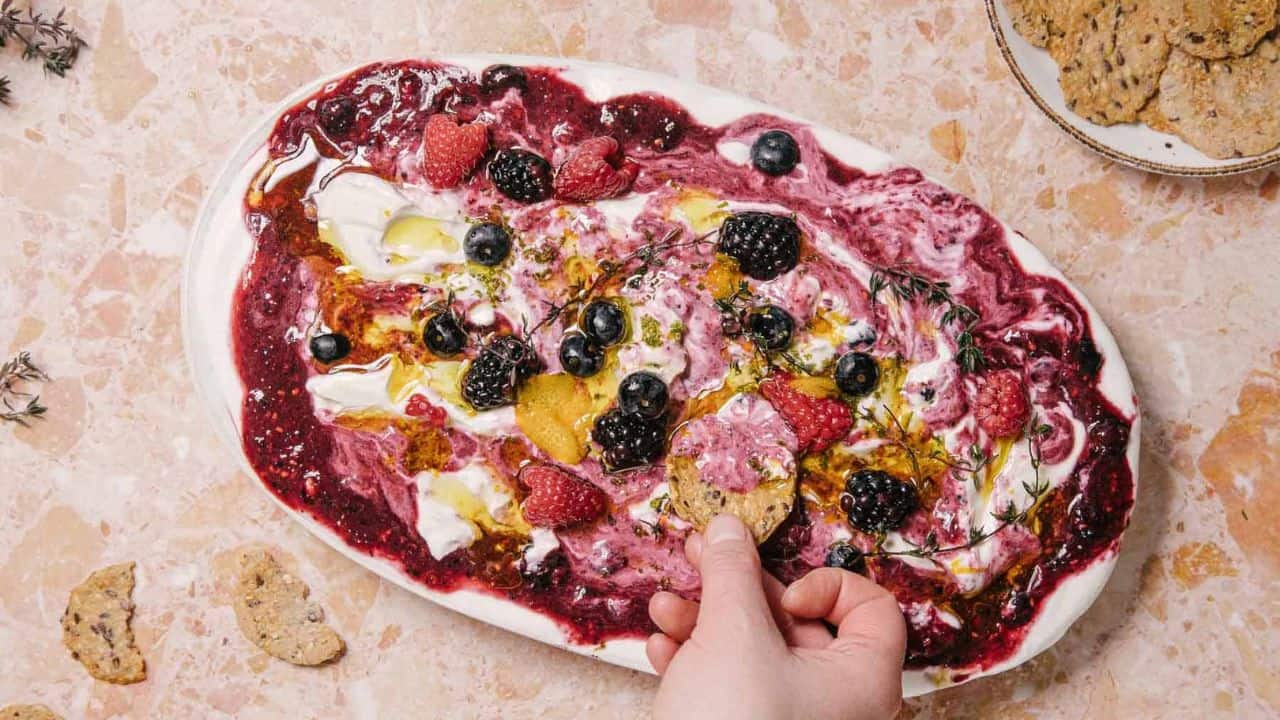 A hand dips a piece of bread into a colorful yogurt dip on an oval platter. The dip features swirls of deep purple and white, topped with raspberries, blueberries, blackberries, and herbs, set on a marbled surface.