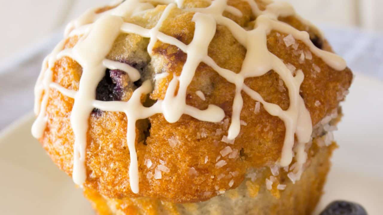 A close-up of a blueberry muffin topped with white icing drizzle and coarse sugar on a white plate.