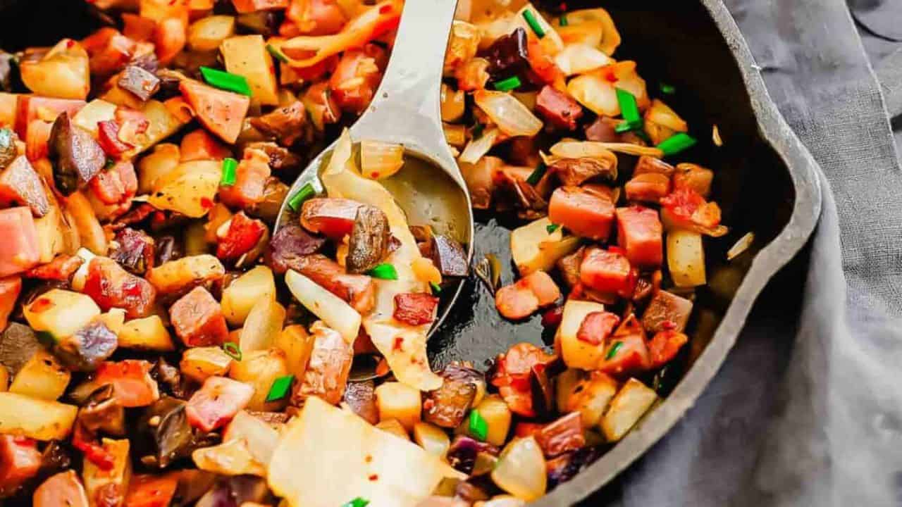 A skillet filled with diced potatoes, onions, and cubed meat, garnished with chopped green herbs, with a metal spoon resting inside.