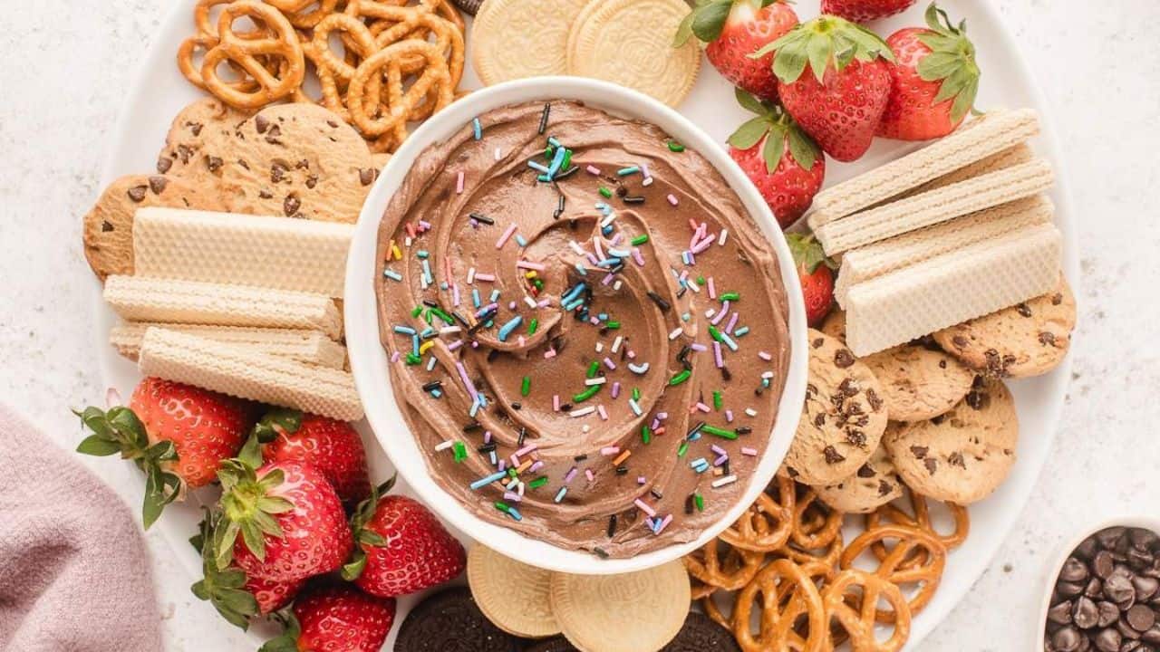 A plate with chocolate dip topped with sprinkles in the center, surrounded by strawberries, chocolate chip cookies, wafer cookies, pretzels, and sandwich cookies.