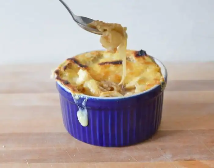 Image shows a Spoon pulling cheese from a bowl of French onion soup in a blue ramekin.