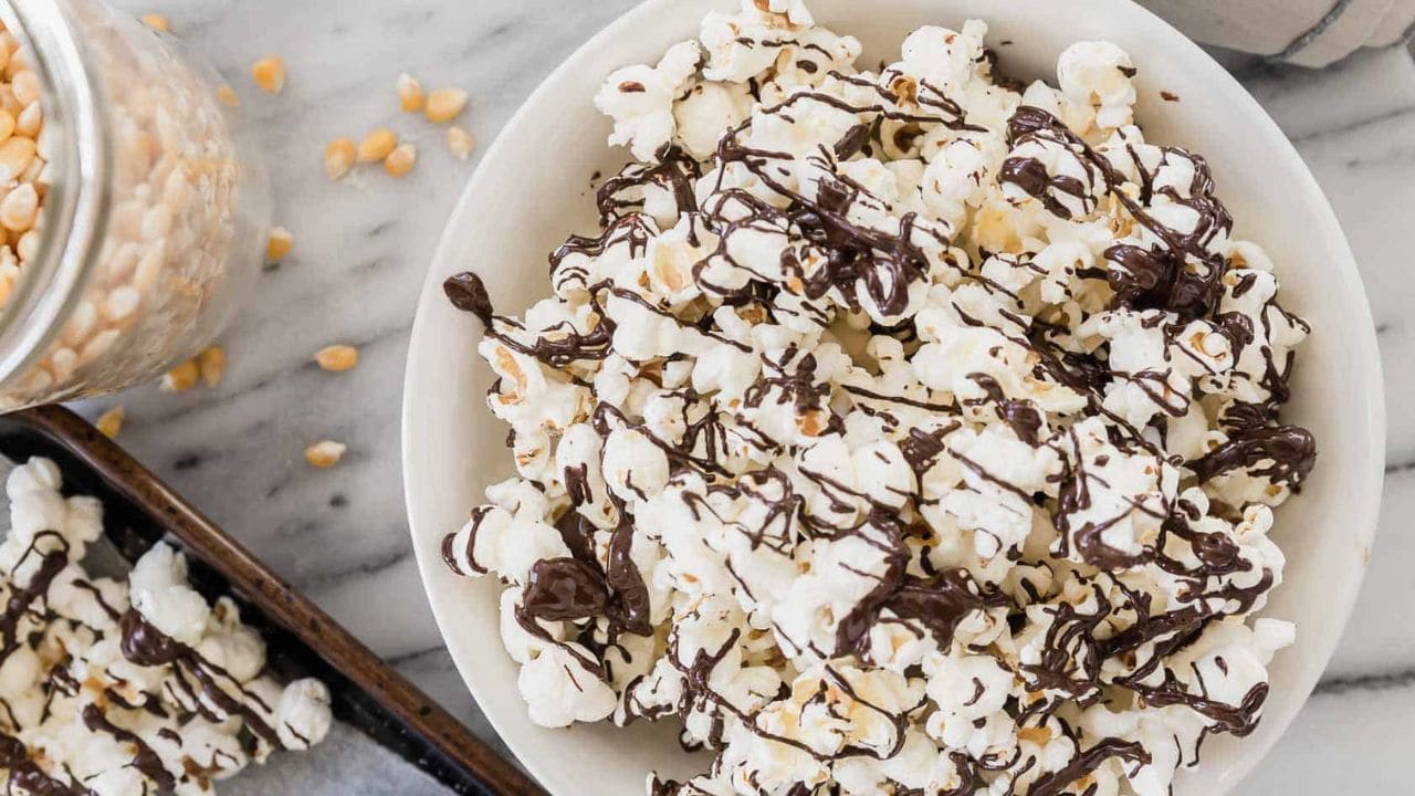 A white bowl filled with popcorn drizzled with chocolate sits on a marble surface, with a baking tray and a jar of popcorn kernels nearby.