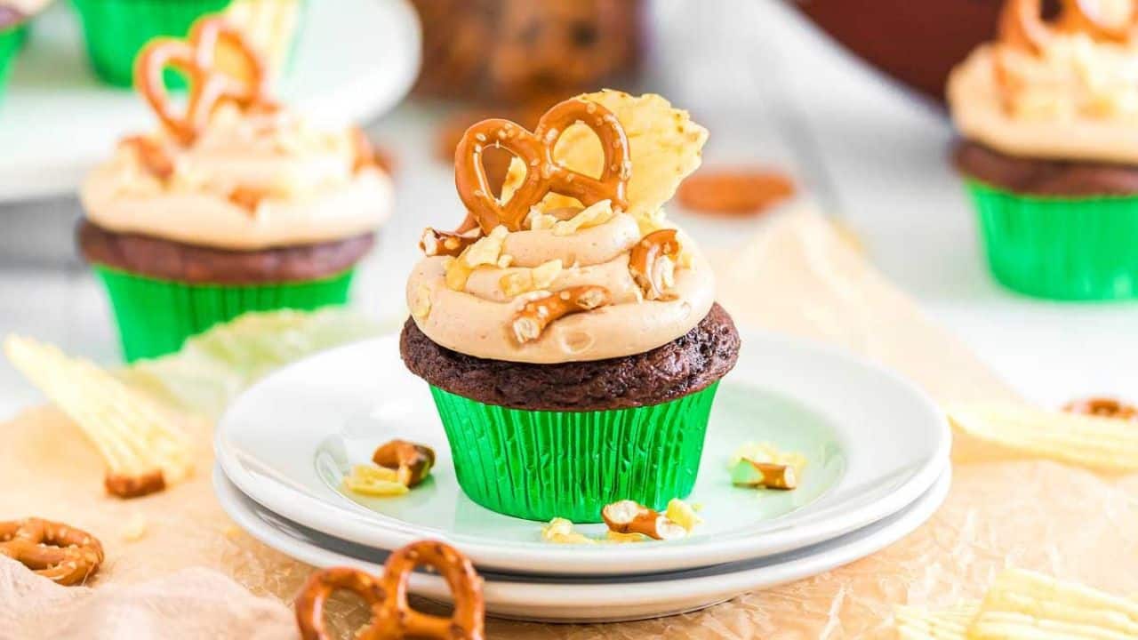 Chocolate cupcake in a green wrapper topped with beige frosting, a pretzel, and a potato chip, placed on a white plate with more cupcakes in the background.