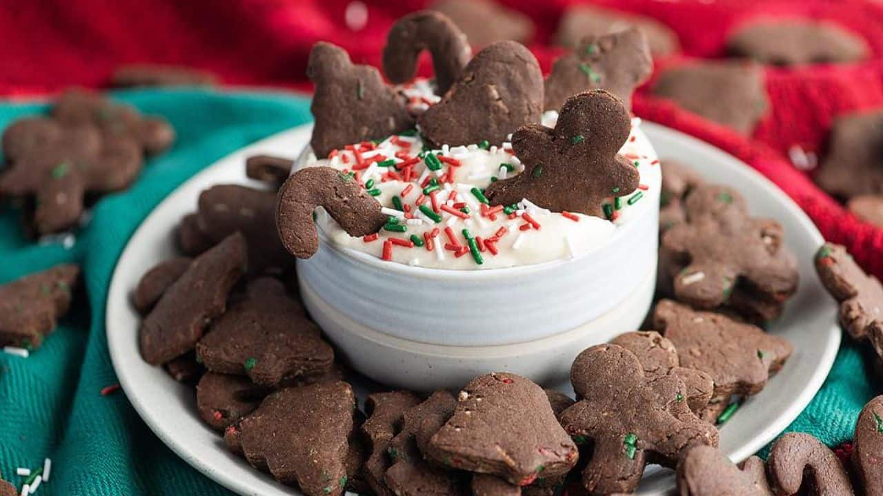A bowl of white dip with red and green sprinkles is surrounded by chocolate holiday cookies shaped like gingerbread men, candy canes, and trees on a plate.