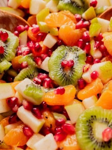 A bowl of fruit salad with sliced kiwi, mandarin orange segments, diced apples, and pomegranate seeds, served with wooden salad utensils.