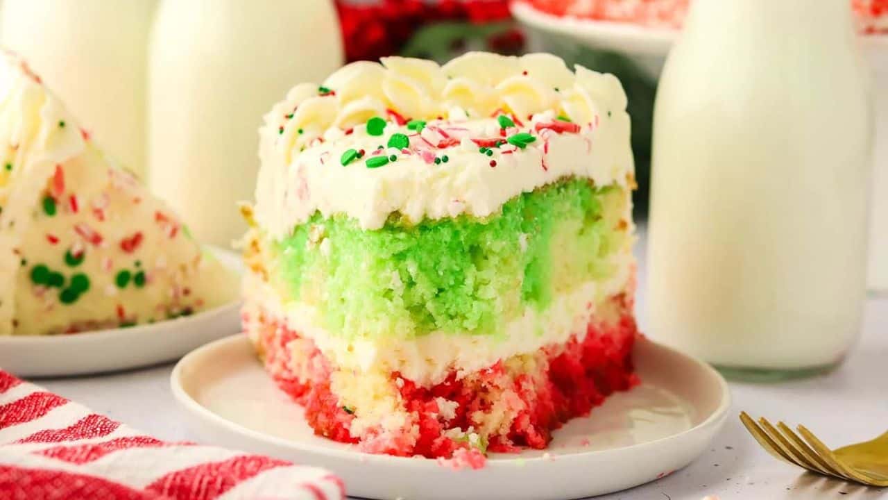 A slice of festive red, green, and white layer cake with white frosting and sprinkles on a plate, next to a bottle of milk.
