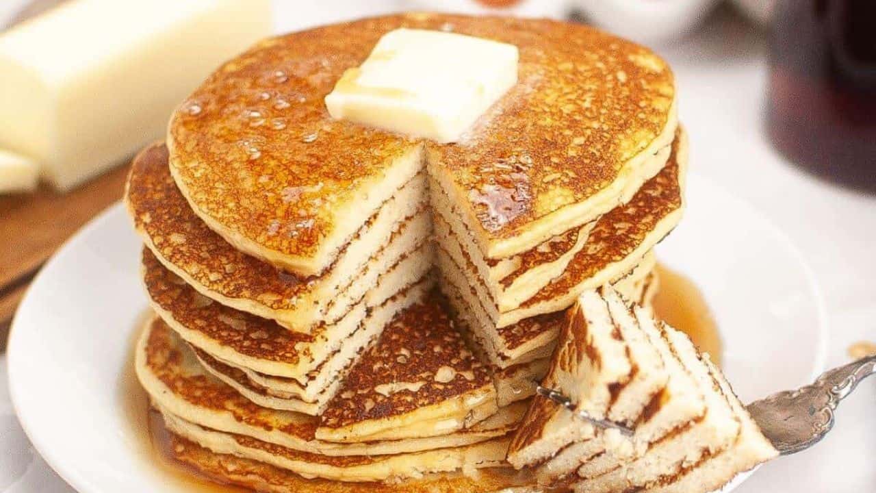 A stack of pancakes with a pat of butter on top, drizzled with syrup. A wedge has been cut out and is resting on a fork beside the stack.