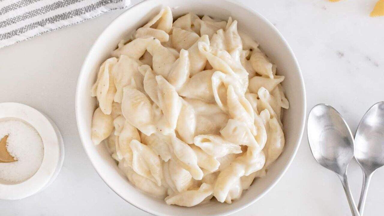 A white bowl filled with creamy shell pasta sits on a white surface next to two metal spoons, a salt container, and a striped cloth napkin.