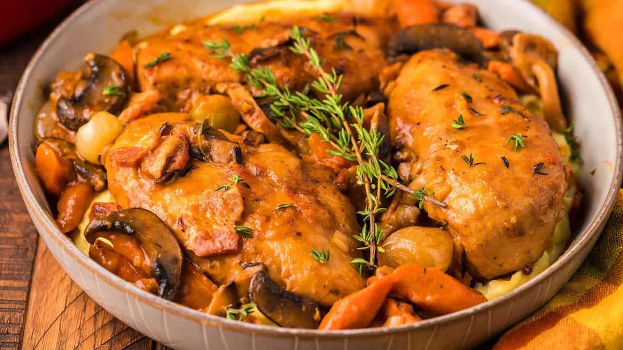 A bowl of cooked chicken breasts with mushrooms, carrots, onions, bacon, and fresh thyme, served over mashed potatoes.