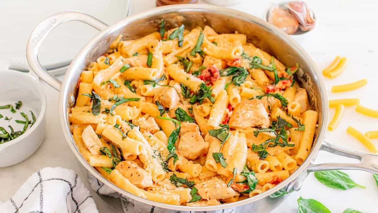 A stainless steel pan filled with creamy pasta, chunks of chicken, spinach, and sun-dried tomatoes, garnished with fresh basil.