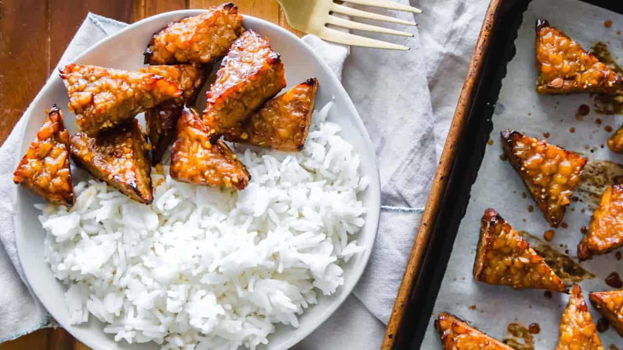 A plate with white rice and glazed triangular pieces of tempeh. More tempeh pieces are on a parchment-lined baking tray beside the plate. A fork is placed nearby.