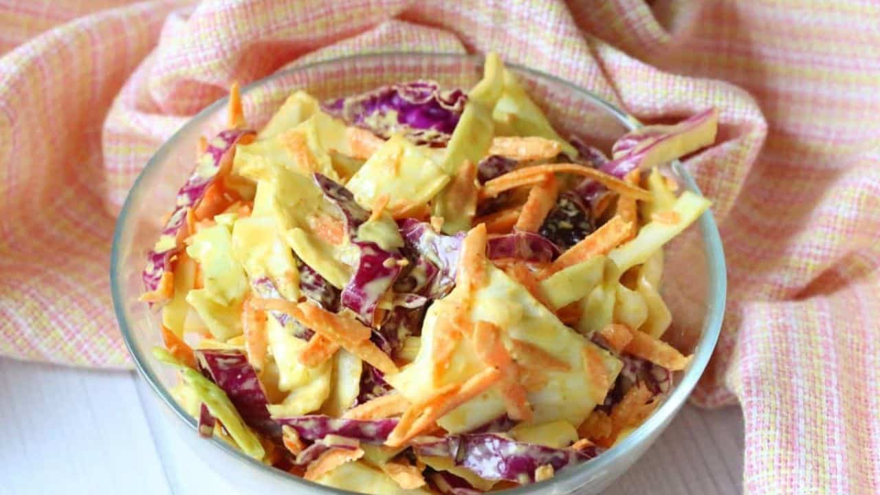 A glass bowl filled with coleslaw made of shredded red and green cabbage and carrots, placed on a pink checkered cloth.