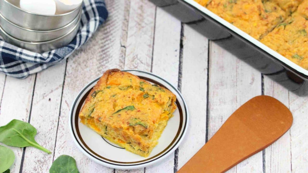 A slice of vegetable casserole on a plate next to a baking dish, wooden spatula, spinach leaves, and stacked metal measuring cups with eggs.