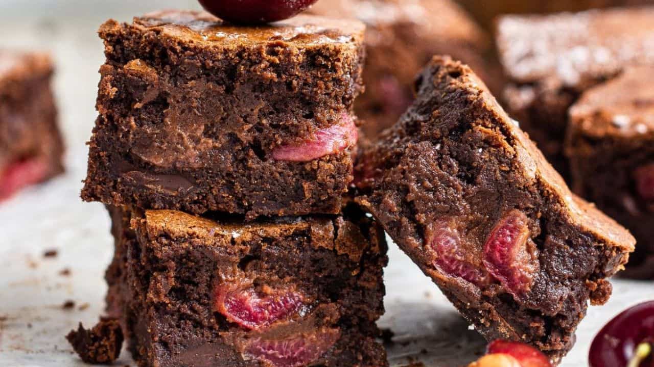 Three stacked chocolate brownies with visible cherry pieces, surrounded by more brownies and fresh cherries.