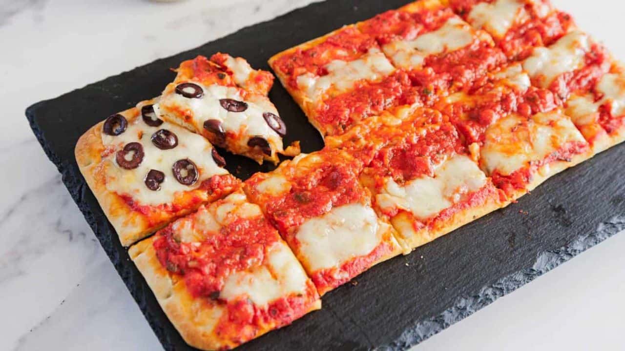 Rectangular pizza cut into square slices on a black slate board, mostly topped with tomato sauce and melted cheese, with one corner slice topped with black olives.