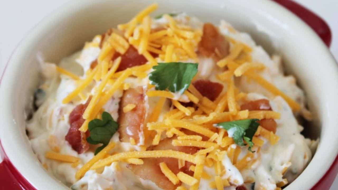 A bowl of creamy dip topped with shredded cheddar cheese, bacon pieces, and fresh parsley.