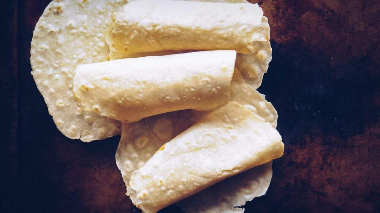 Three rolled corn tortillas rest on top of two flat corn tortillas, all on a dark brown surface.