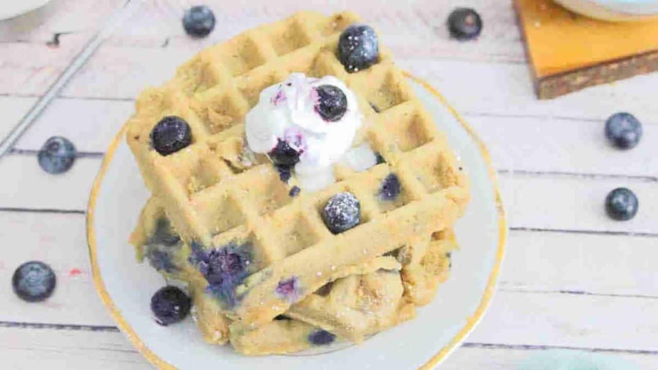 A stack of blueberry waffles topped with whipped cream and fresh blueberries sits on a white plate.