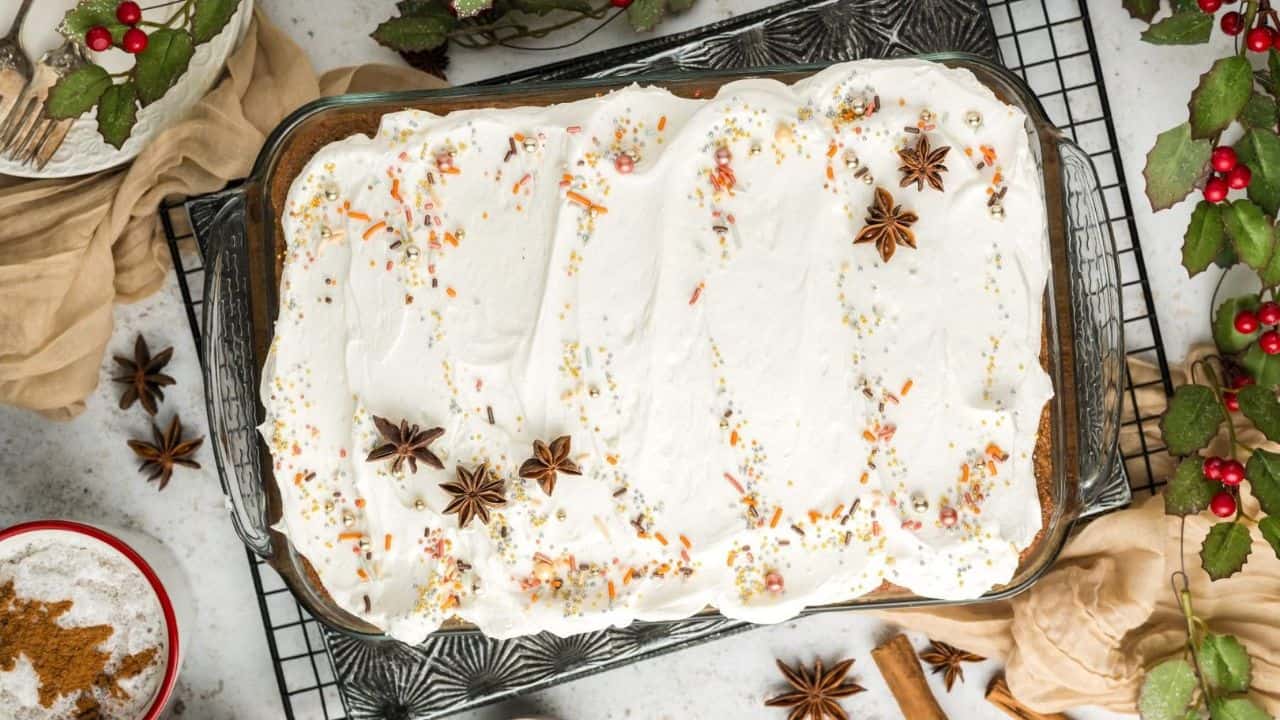 A glass baking dish filled with cake topped with white frosting, star anise, and sprinkles, shown on a cooling rack surrounded by holiday decor.