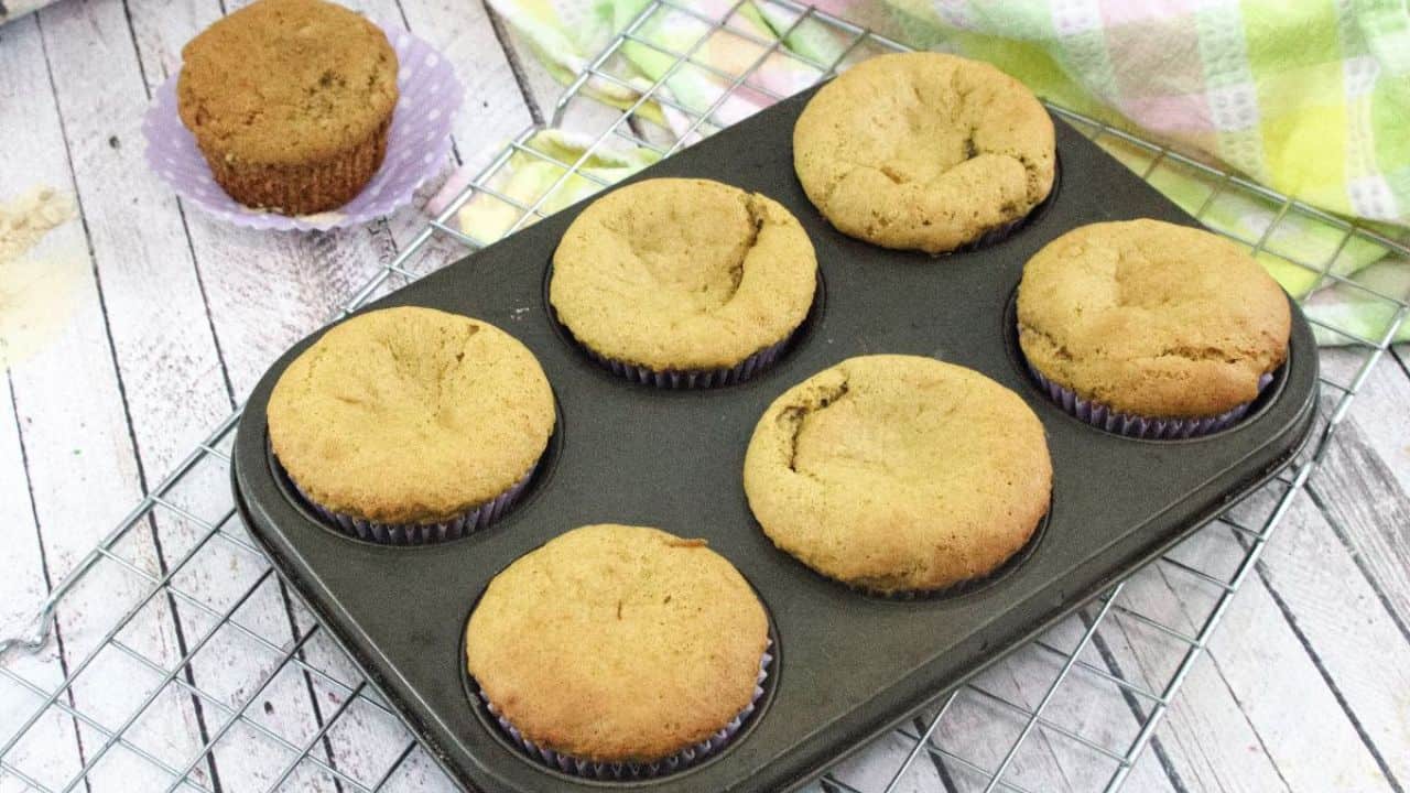Six baked muffins sit in a muffin tray on a wire cooling rack, with one muffin placed separately in a purple wrapper beside the tray.