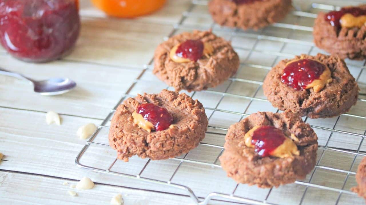 Chocolate cookies with peanut butter and jelly centers cooling on a wire rack, with a jar of red jelly and a spoon in the background.