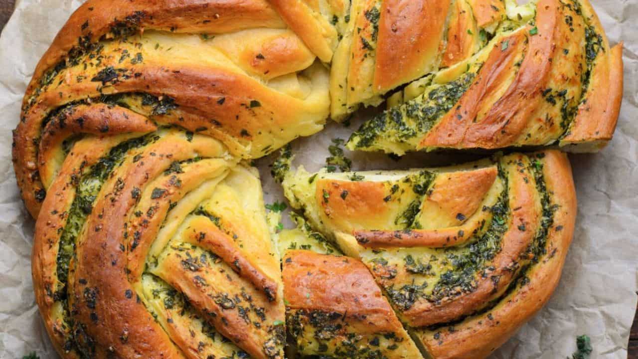 A round, braided bread with a golden crust, swirled with green herbs, sits on parchment paper. Some slices have been cut and partially pulled out.