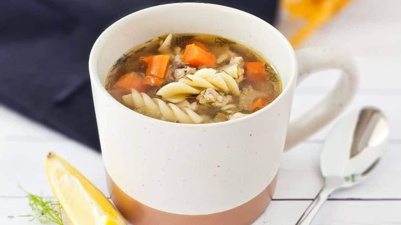 A white mug filled with soup containing spiral pasta, diced carrots, and herbs sits on a white surface next to a spoon and a lemon wedge.