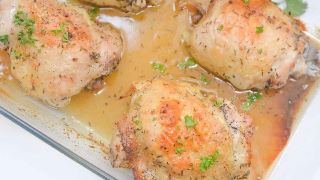 Baked chicken thighs with golden, crispy skin in a glass baking dish, garnished with chopped parsley and surrounded by pan juices.