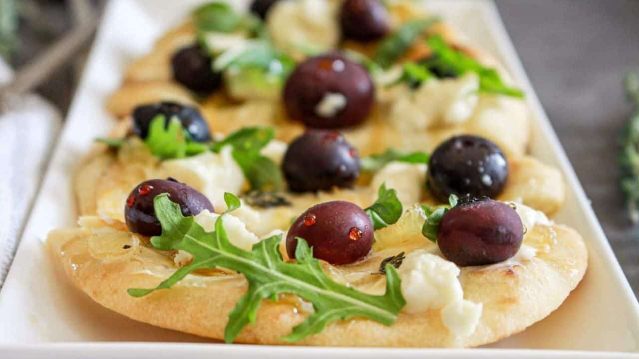 A close-up of a goat cheese flatbreads topped with caramelized onions, arugula, and black blueberries, served on a white plate.