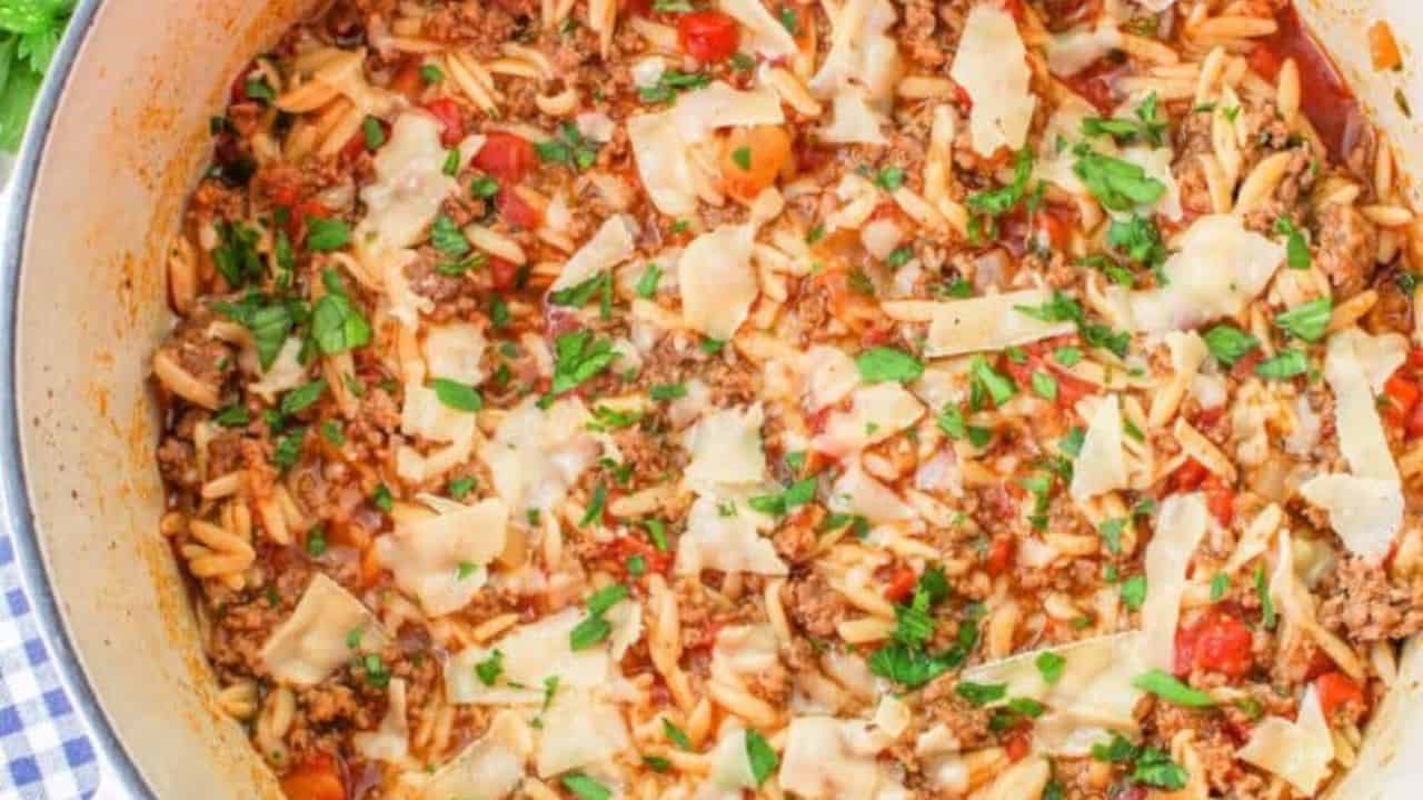 A plate of pasta with minced meat, cherry tomatoes, and cheese shavings on a white plate. A fork rests on the plate, and a blue checkered napkin is underneath. Parsley is visible beside the plate.