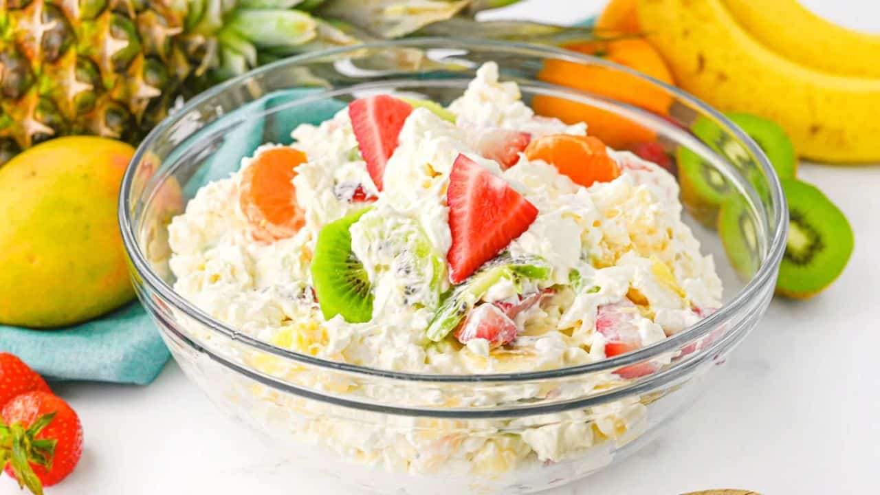 A glass bowl of creamy fruit salad with sliced strawberries, kiwi, mandarin oranges, and pineapple, surrounded by whole fruits on a table.