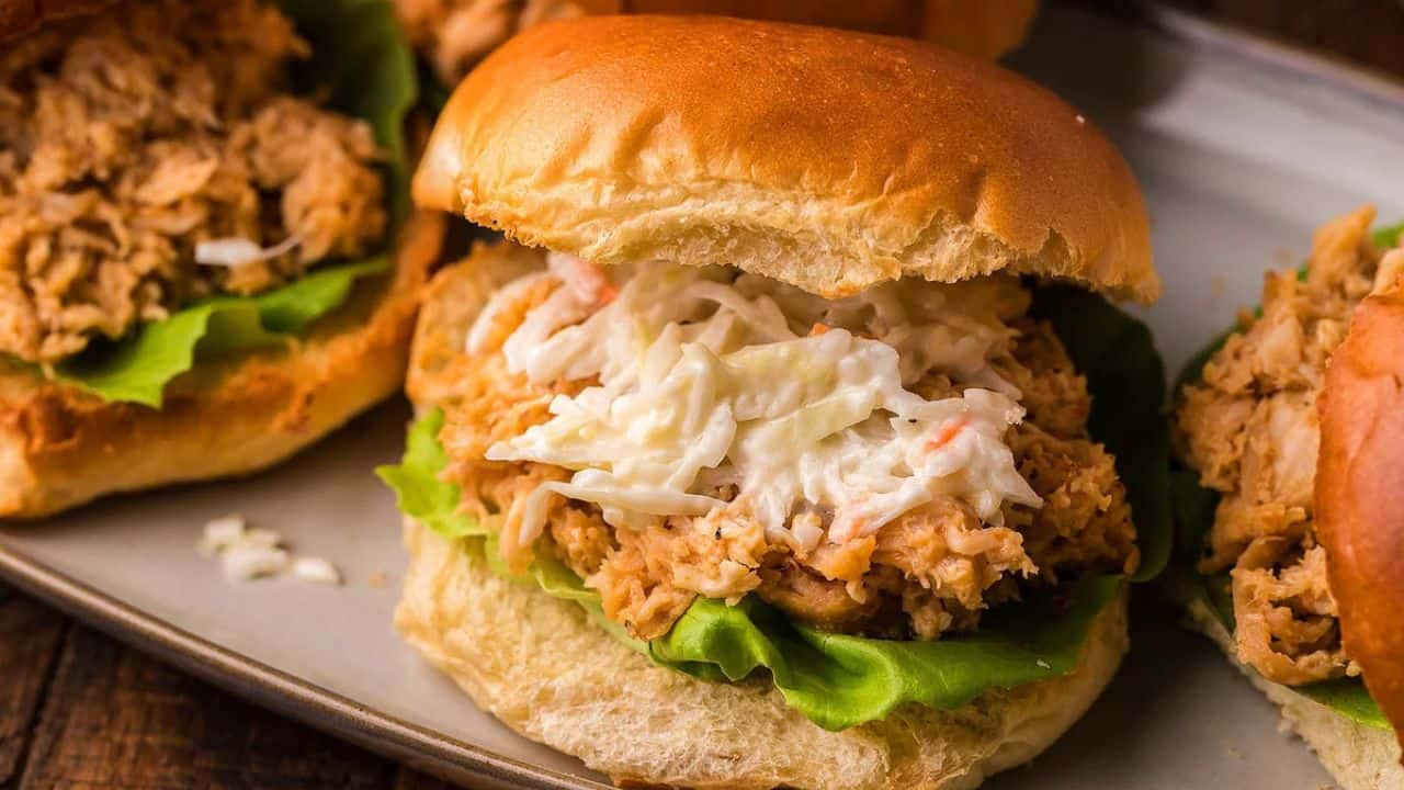 A sandwich with shredded chicken, lettuce, and coleslaw on a bun sits on a plate with other similar sandwiches in the background.