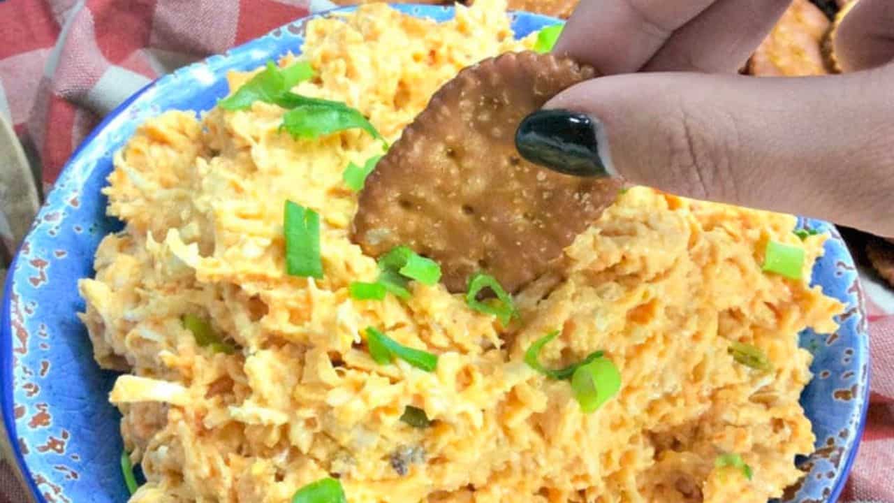 A hand dips a round cracker into a bowl of shredded cheese dip topped with sliced green onions.