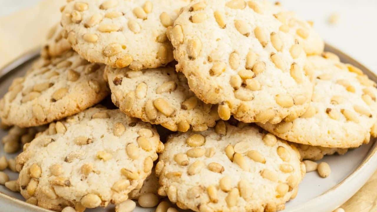 A plate stacked with round cookies topped with pine nuts, with some pine nuts scattered on the plate.