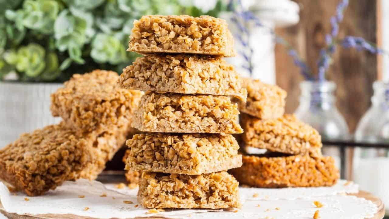 A stack of oat flapjacks sits on parchment paper with more flapjacks and green plants visible in the background.
